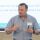 A man in a blue striped shirt speaks while holding a clicker. A screen behind him displays a quote starting with "We exist to make the workplace a more fulfilling place...