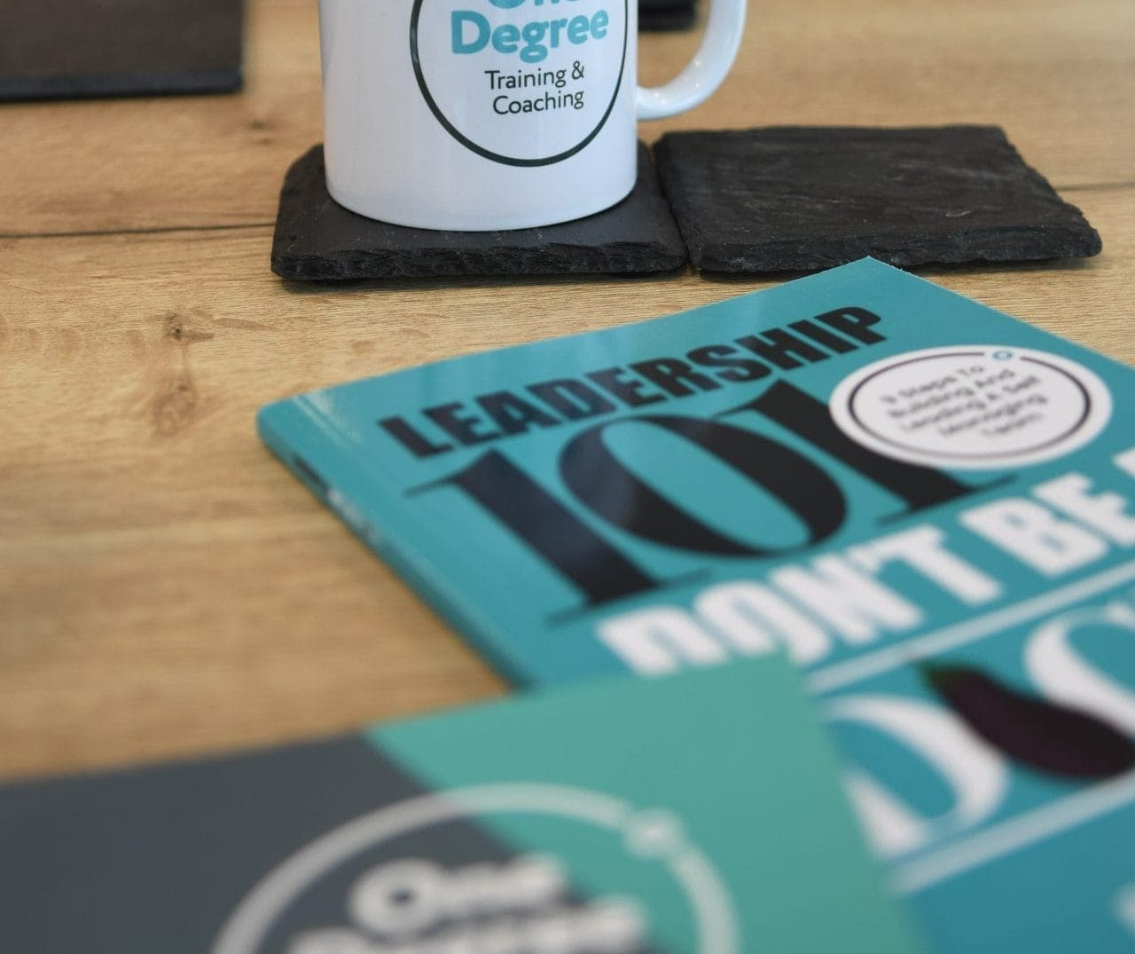 A white mug with "One Degree Training & Coaching" on it sits on a wooden table, surrounded by leadership books and a potted plant in the background.