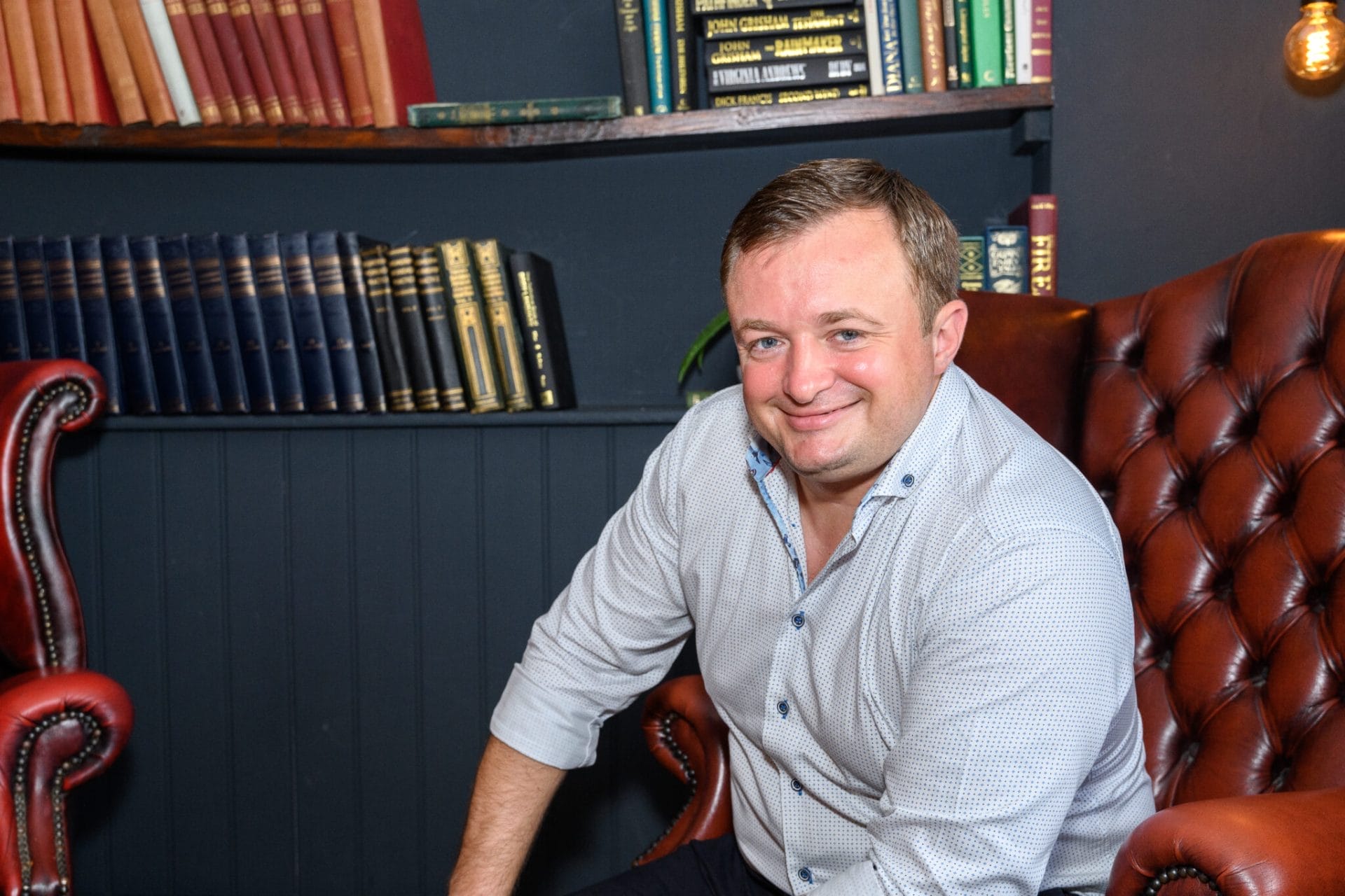 A man in a light blue button-down shirt sits in a brown leather armchair in a room with shelves filled with books.