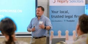 A man in a blue shirt, recently awarded Managing Director of the Year, presents in front of a screen slide and an advertisement for Hegarty Solicitors. Several people are seated and listening attentively to the presentation.