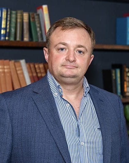 Man in a blue blazer and striped shirt standing in front of a bookshelf filled with various books.