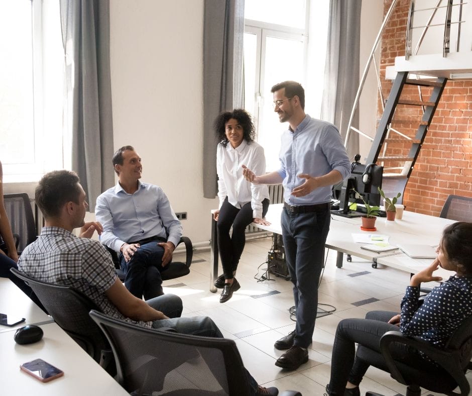 A group of people sit and stand in an office space, engaged in a discussion, providing examples of situational leadership in action.