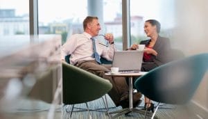 Two people in business attire sit comfortably in an office lounge area, smiling and talking while holding coffee cups. A laptop is on the small table between them.