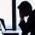 A person in a suit sits at a desk with their head in their hands in front of a laptop, silhouetted against a large window.